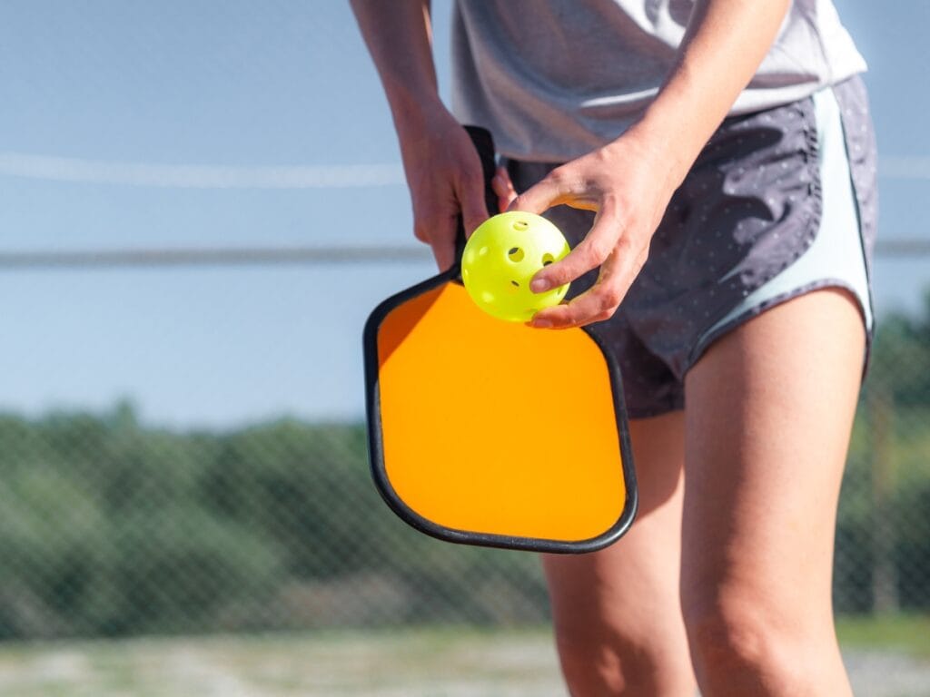 A person holding a yellow pickleball and an orange paddle on an outdoor court, preparing to serve, highlights the unique gear used in pickleball vs tennis.