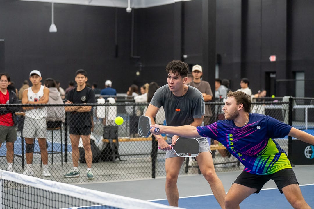Two men play pickleball indoors, focusing on the ball as one lunges to hit it. Behind a chain-link fence, spectators watch the action, highlighting the energy of Pickleball vs Tennis matches.