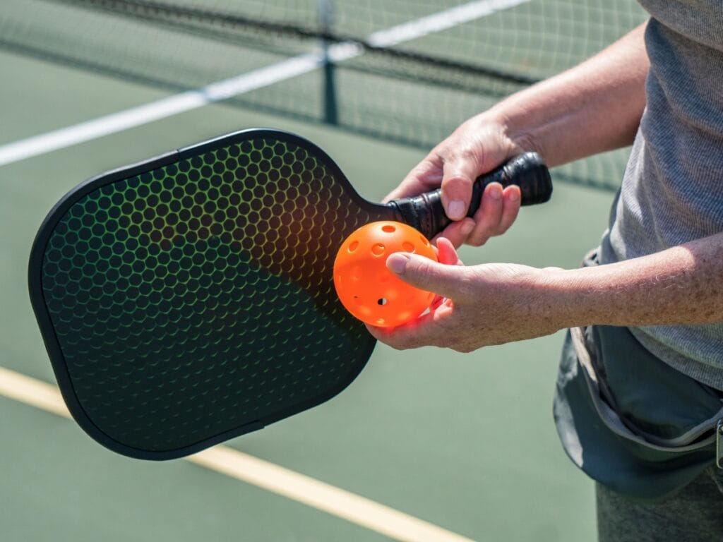 A person displays the skills of pickleball, holding a black pickleball paddle and an orange perforated ball on an outdoor court.
