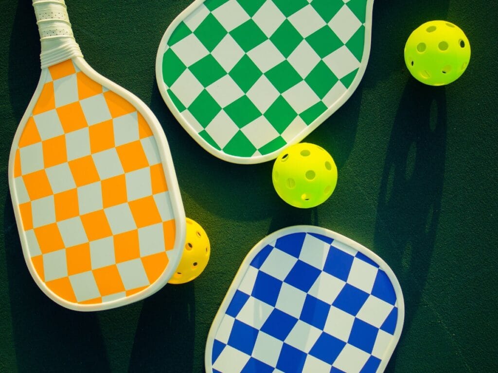Three pickleball paddles with checkered patterns in orange, green, and blue—plus four yellow pickleballs—are arranged on a dark surface, showcasing some of the essential equipment needed for pickleball.