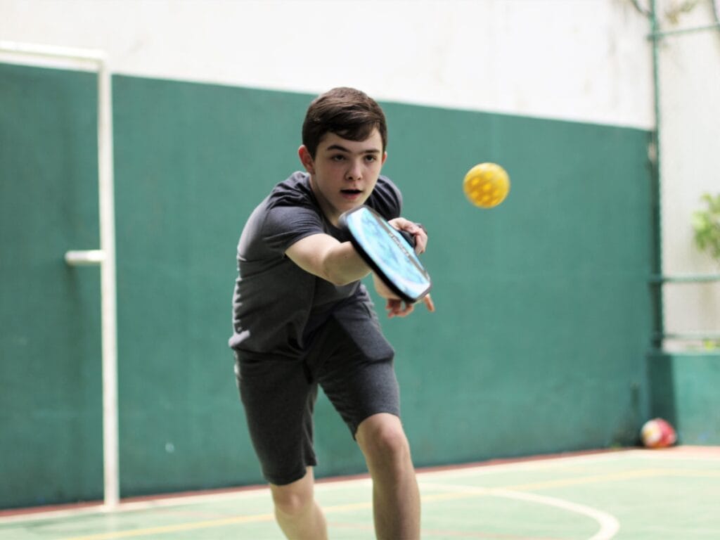 A boy in athletic clothing plays pickleball indoors, reaching forward with a paddle—essential equipment needed for pickleball—to hit a yellow ball.