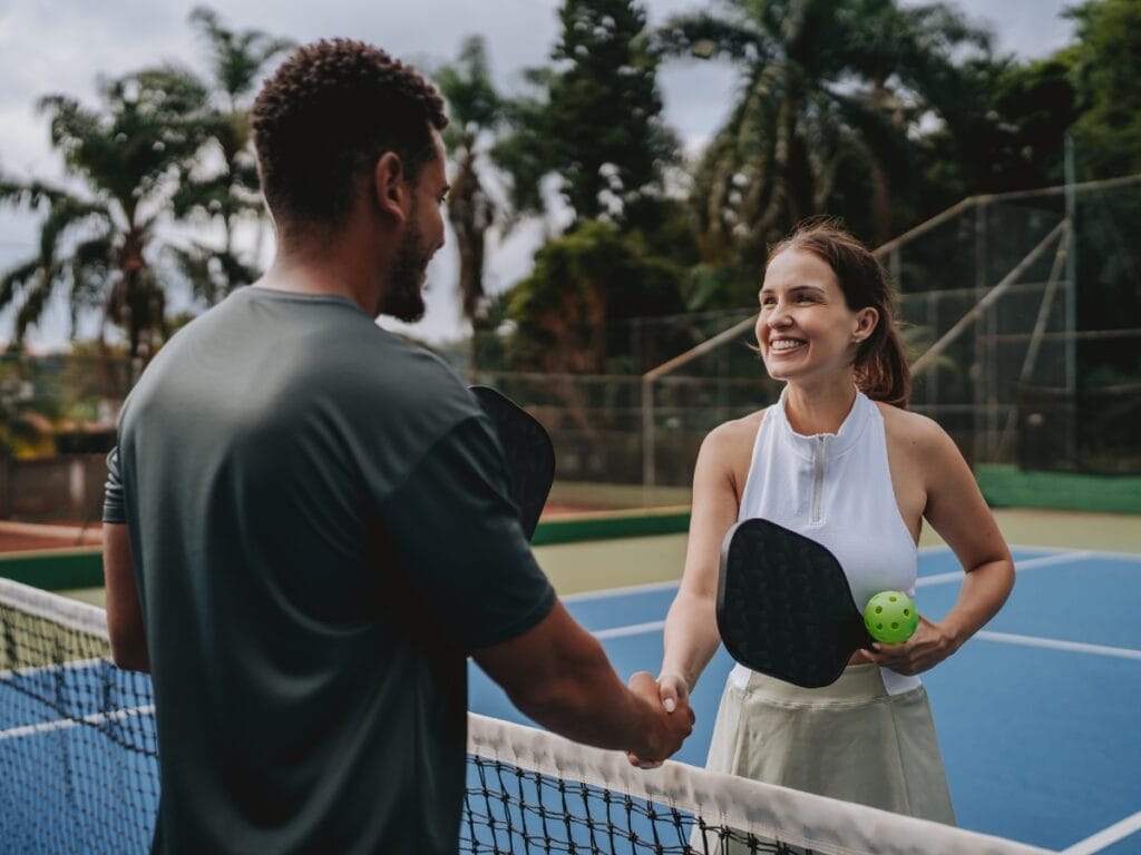 Two people stand on a pickleball court, smiling as they shake hands over the net after a friendly pickleball match; the woman holds a paddle and ball.