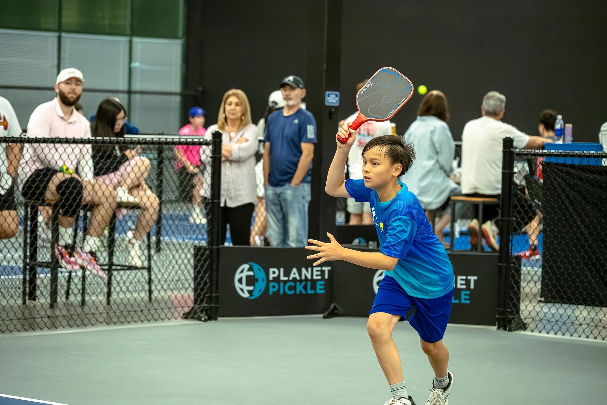 A young boy in blue sportswear prepares to hit a ball with a paddle on an indoor pickleball court, showcasing key pickleball gear as spectators watch behind a fence.