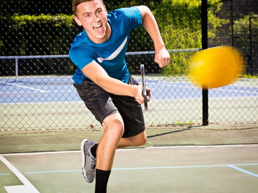 A young man in athletic attire lunges forward to hit a yellow pickleball with a paddle on an outdoor court, showcasing one of the effective pickleball strategies for beginners.