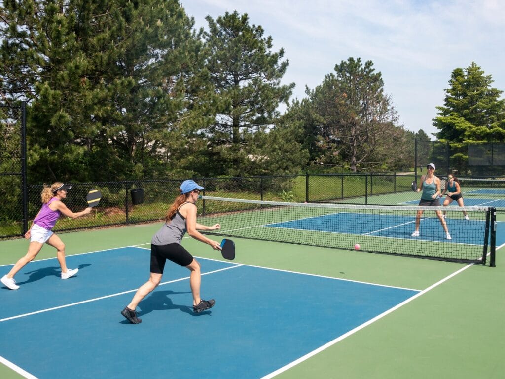 Four women play a doubles pickleball match on an outdoor court surrounded by trees under a clear sky, putting Pickleball Strategies for Beginners into practice as they enjoy the game.