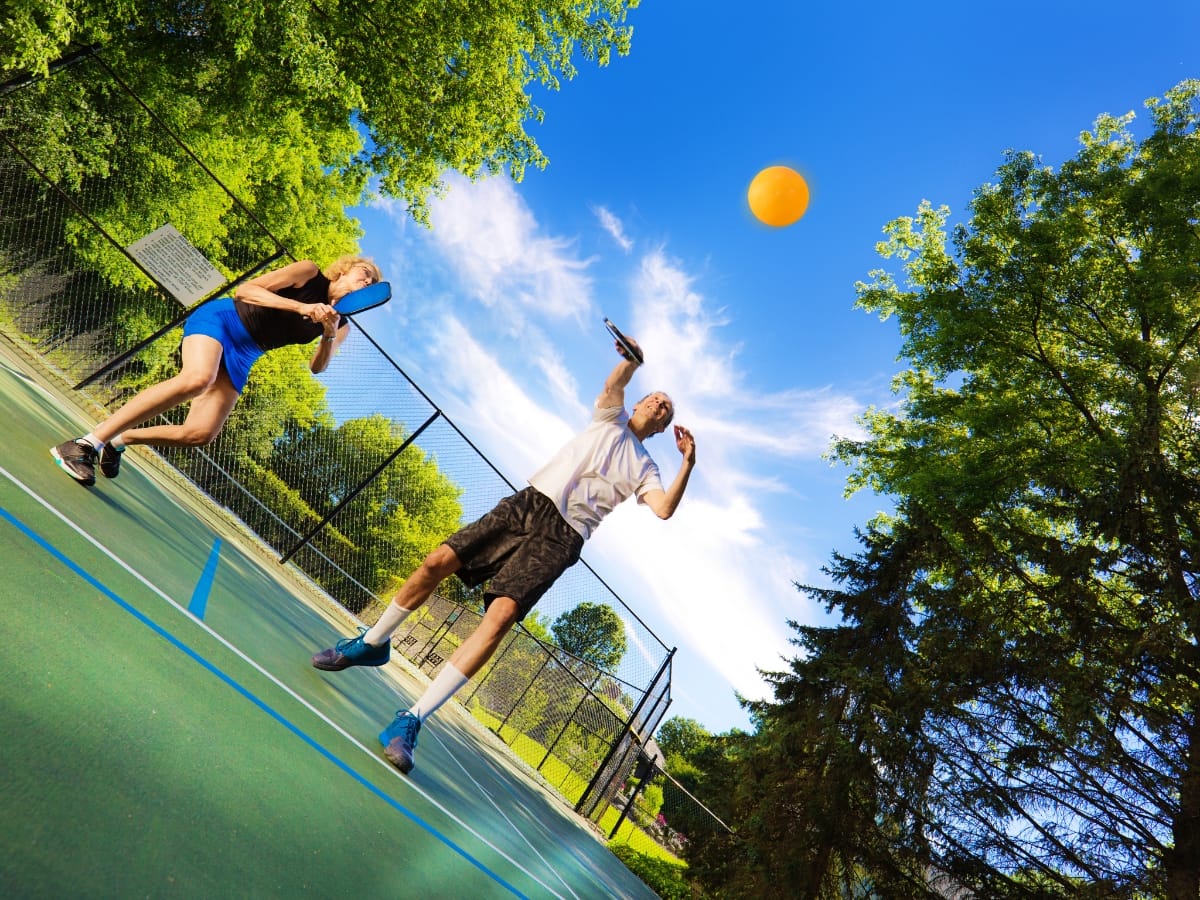 Two people are playing pickleball on an outdoor court, both holding paddles and watching a yellow ball in the air under a clear blue sky as they practice pickleball strategies for beginners.