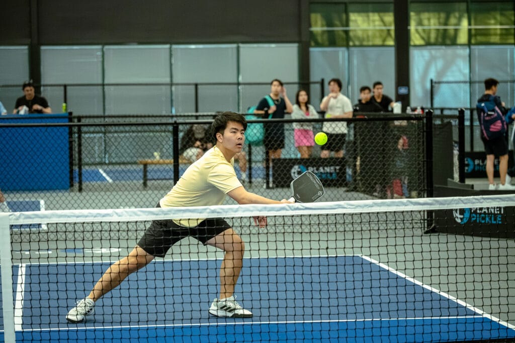 Top Pickleball Injuries: How to Avoid Them and Play Safely 9 A man in athletic wear plays pickleball indoors, preparing to hit a ball with a paddle as spectators watch from behind a fence, demonstrating how to play pickleball safely and avoid pickleball injuries.