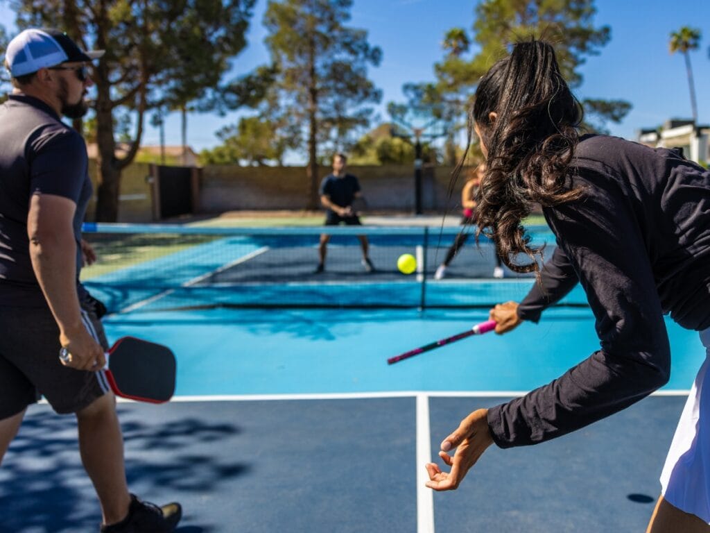 Four people play pickleball on an outdoor court, with one woman in the foreground preparing to hit the ball—demonstrating why proper technique is important to avoid top pickleball injuries. Trees and a fence are visible in the background.