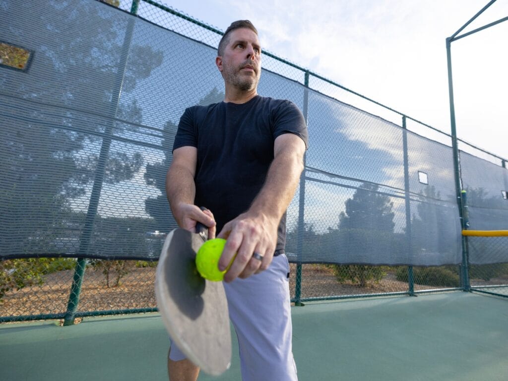 A man in a black shirt and light shorts holds a paddle and yellow ball on an outdoor sports court, preparing to serve—demonstrating proper form to help prevent top pickleball injuries.