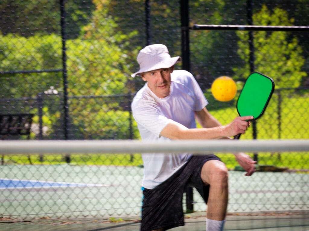 An older man wearing a white hat and shirt plays pickleball on an outdoor court, focused on avoiding pickleball faults as he prepares to hit a yellow ball with his green paddle.