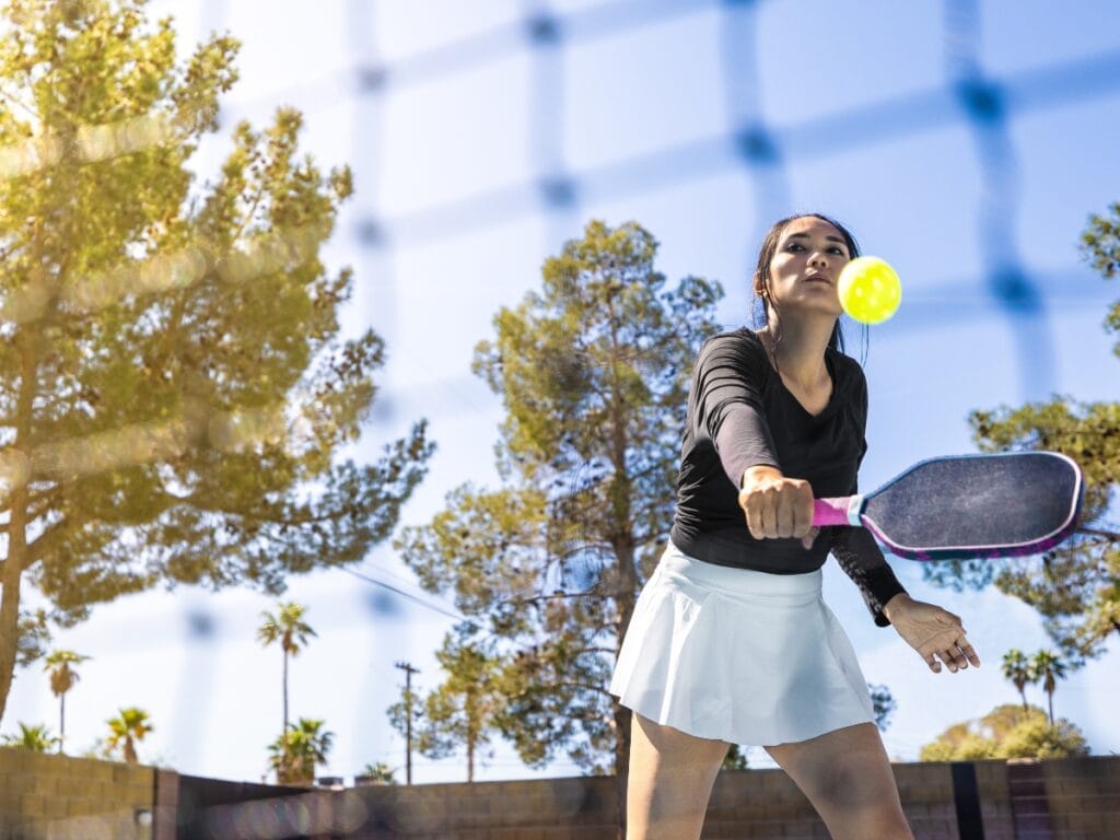 A woman in athletic attire returns a ball with a paddle on an outdoor pickleball court, avoiding common pickleball faults, with trees and blue sky in the background.