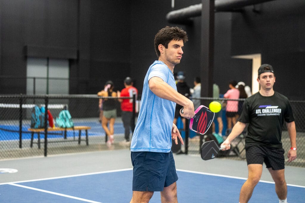Common Pickleball Faults Explained 8 Two men play pickleball indoors—one in a light blue shirt hitting the ball with a paddle, the other in black nearby. Others watch in the background, ready to spot any pickleball mistakes or Pickleball faults during their match.