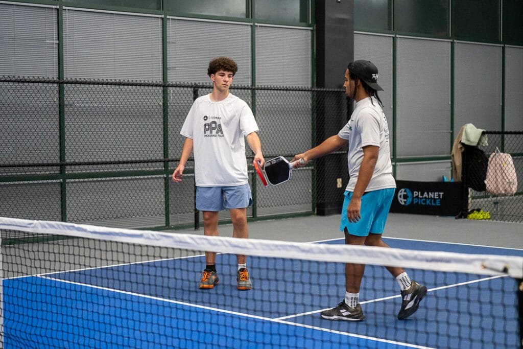 Proper Score Calling Etiquette in Pickleball 7 Two men on an indoor pickleball court reach out to tap paddles over the net after a game, showcasing great sportsmanship and etiquette.