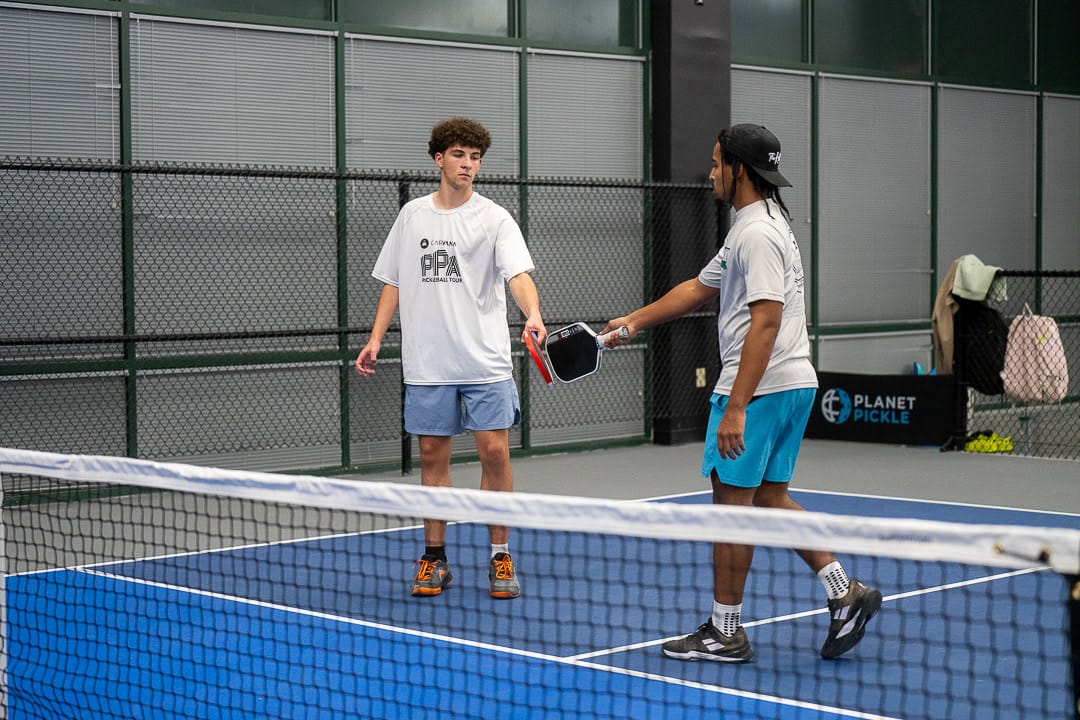 Two men on an indoor pickleball court reach out to tap paddles over the net after a game, showcasing great sportsmanship and etiquette.