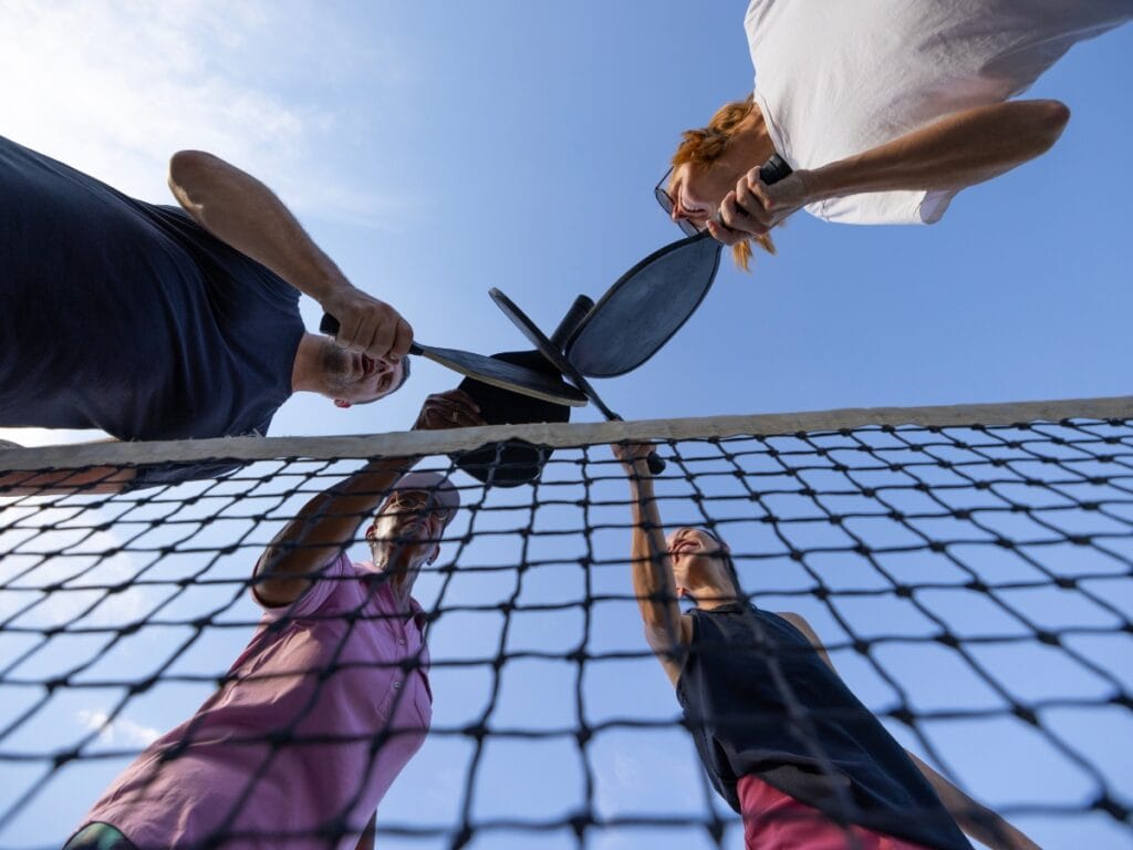 Four people stand around a net, holding pickleball paddles together in the center, viewed from below against a blue sky as they prepare for score calling before the next rally.