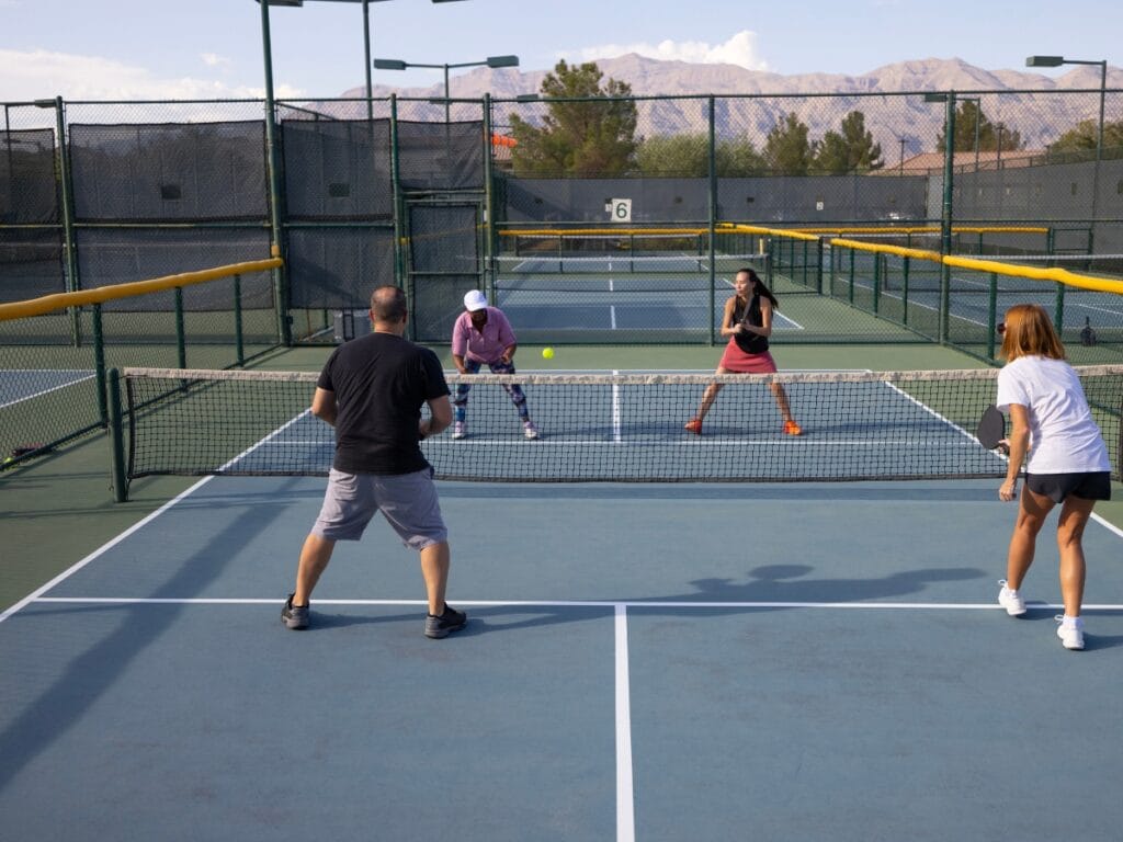 Four people play a doubles pickleball match on an outdoor court with mountains in the background, engaging in energetic rallies and clear score calling.
