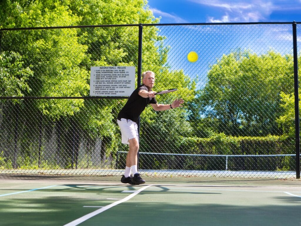 A man in athletic wear prepares to hit a tennis ball with a racket on an outdoor tennis court, ready for score calling, with a sign and trees visible in the background.