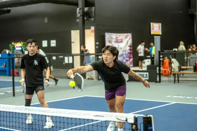 Two people play pickleball indoors; one lunges forward with a paddle to hit the ball while the other stands behind, observing the action&mdash;illustrating how Pickle Coaching can benefit players of all levels.