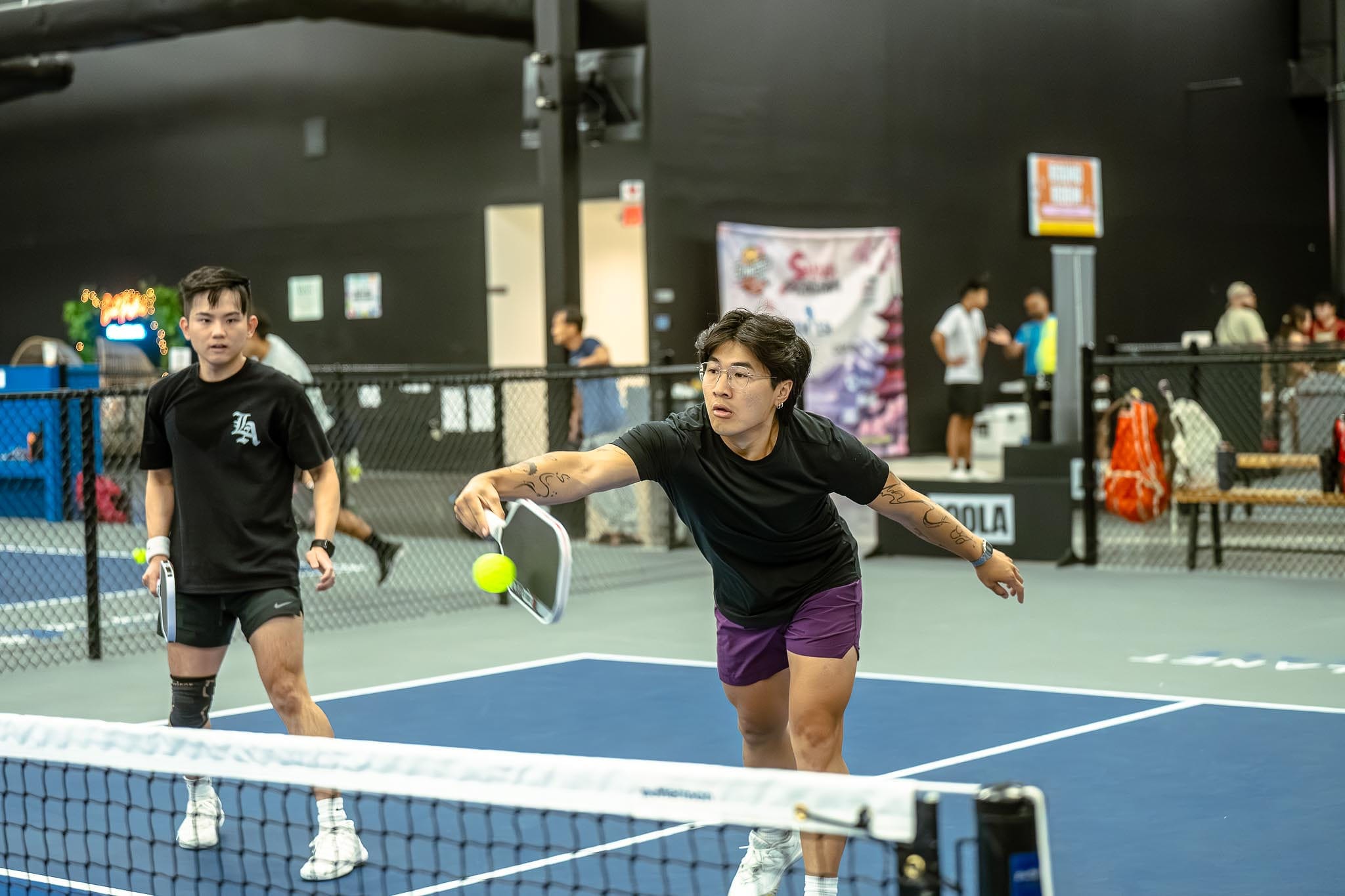 Two people play pickleball indoors; one lunges forward with a paddle to hit the ball while the other stands behind, observing the action—illustrating how Pickle Coaching can benefit players of all levels.