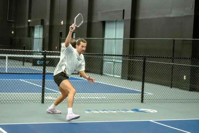 A person in athletic wear, like many pickleball players, prepares to hit a pickleball with a paddle on an indoor court, with a black fence and dark walls in the background.
