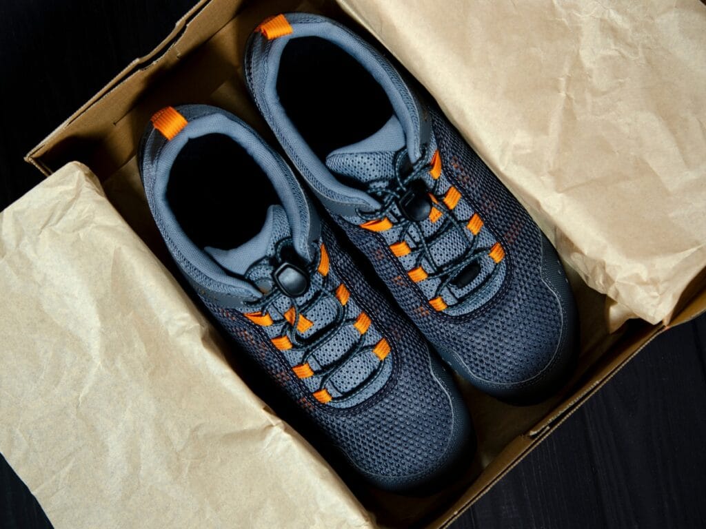 A pair of gray athletic shoes with orange accents is placed inside a cardboard shoe box lined with brown tissue paper, ready to showcase stylish new shoes.