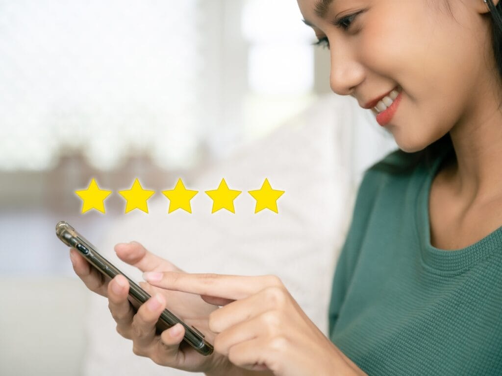 A woman smiles while using a smartphone to leave a five-star review for her new shoes, with five yellow stars displayed, indicating a positive rating.