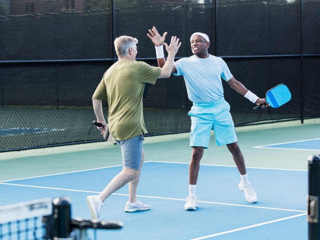 Two men on a pickleball court, both in athletic clothing and sturdy shoes, share a high five. One holds a paddle, and both are smiling.