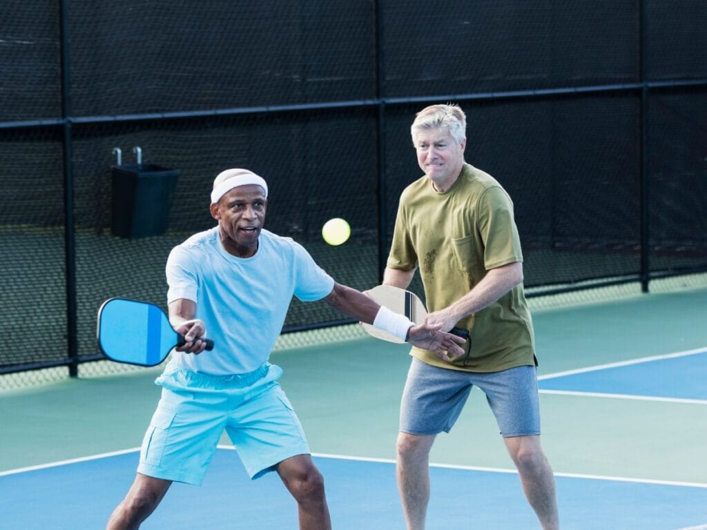 Two seniors play doubles pickleball outdoors; one prepares to hit the ball with his paddle while the other stands behind him watching, highlighting the benefits of staying active together.