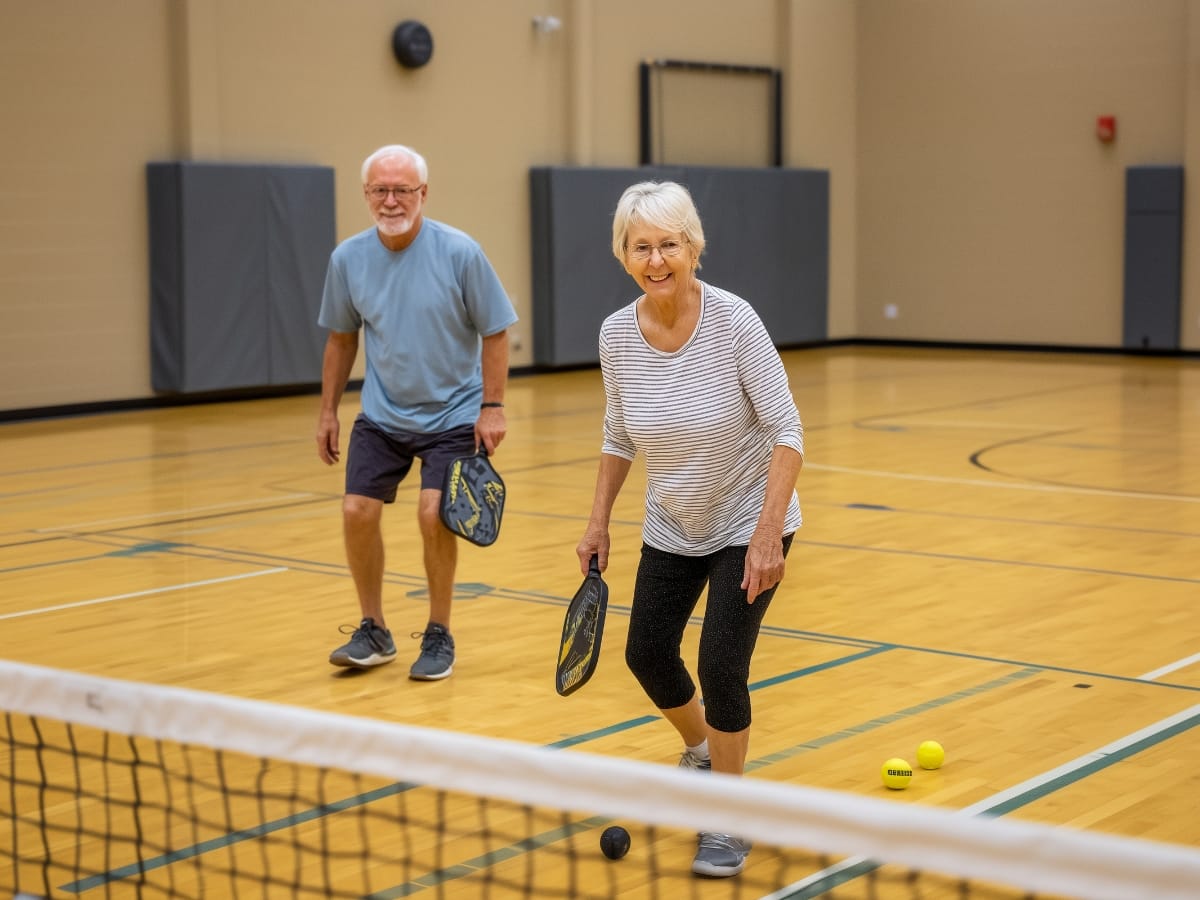 Two older adults play pickleball indoors on a gym court, both holding paddles and smiling, enjoying the benefits of pickleball for senior fitness with pickleballs on the floor near the net.