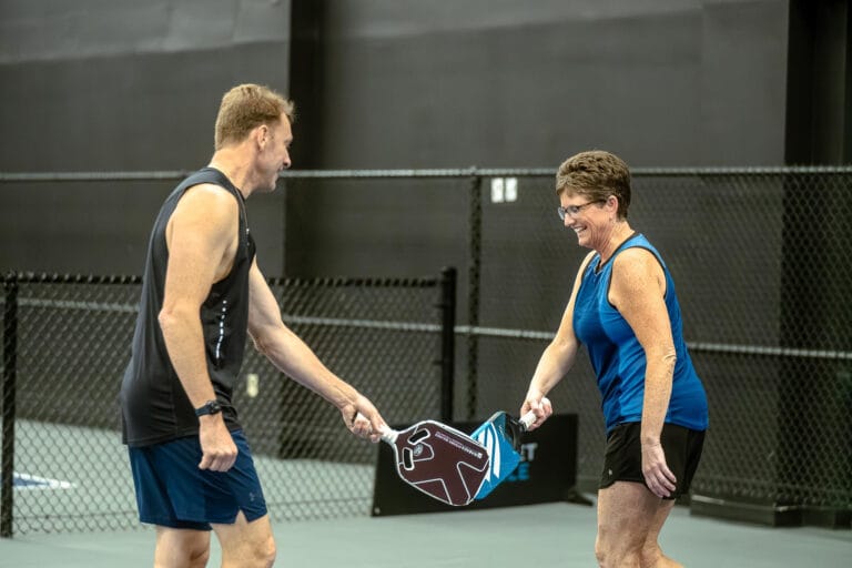 A man and a woman on an indoor court tap pickleball paddles together and smile, standing near a black chain-link fence, enjoying the benefits of staying active as seniors.