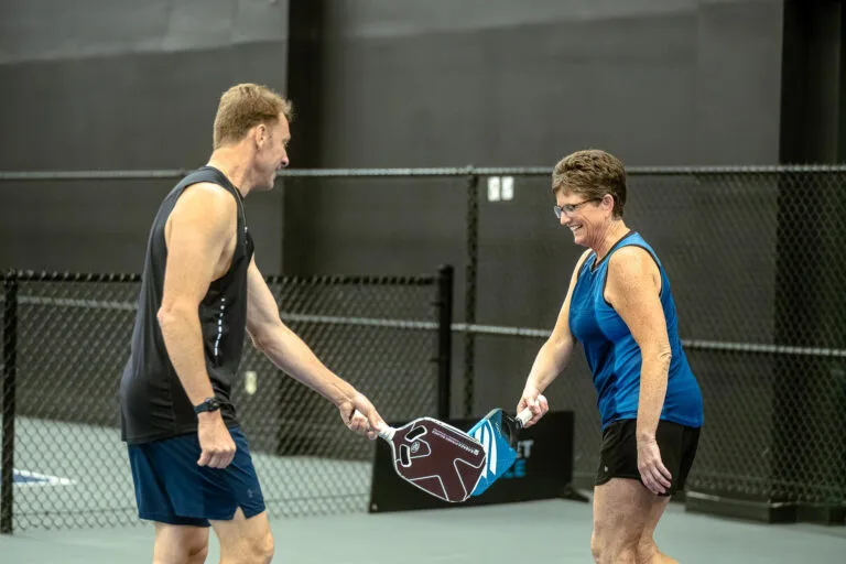 A man and a woman on an indoor court tap pickleball paddles together and smile, standing near a black chain-link fence, enjoying the benefits of staying active as seniors.