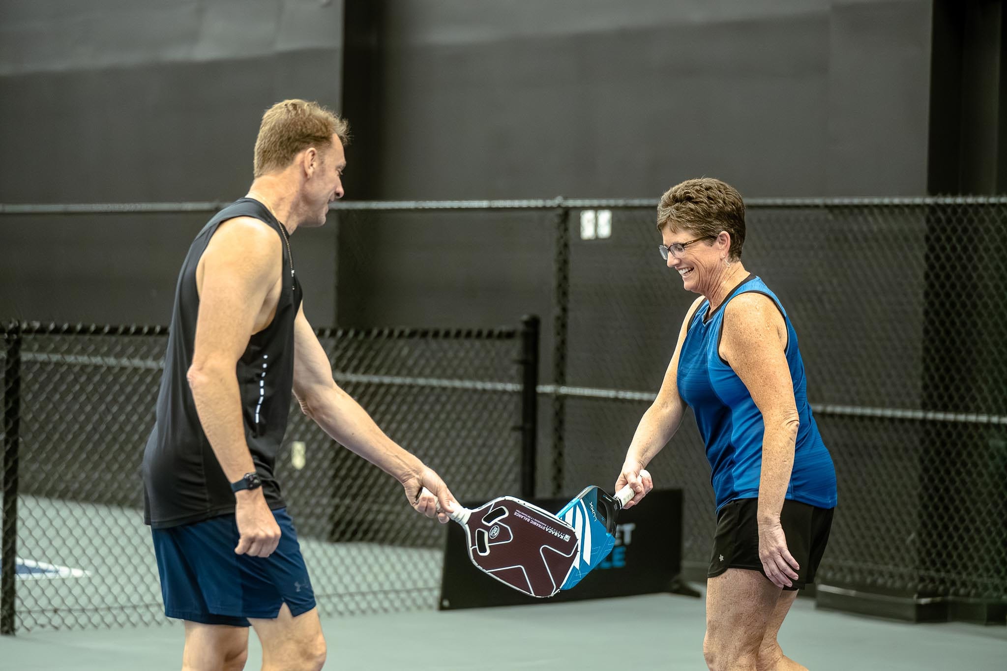 A man and a woman on an indoor court tap pickleball paddles together and smile, standing near a black chain-link fence, enjoying the benefits of staying active as seniors.