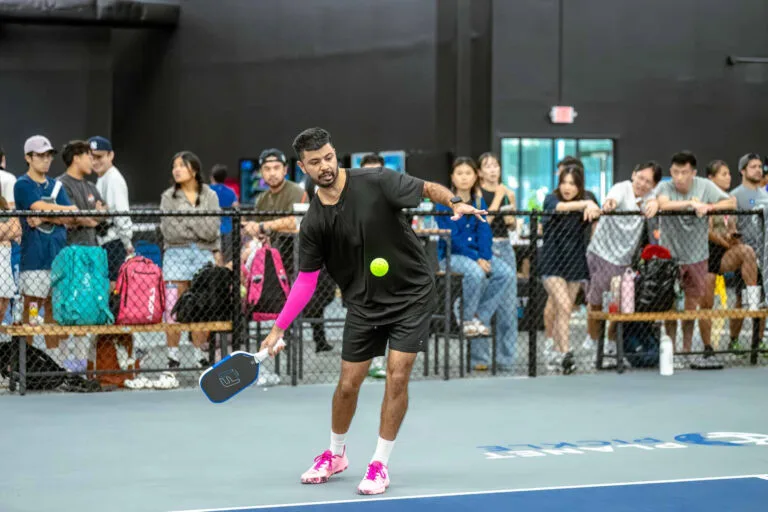 A man in athletic wear follows his essential pre game checklist before playing pickleball indoors, as a crowd watches from behind a fence.