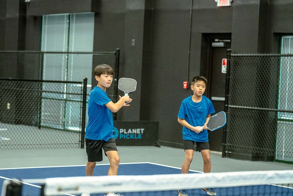 Two boys in blue shirts playing doubles pickleball on an indoor court, both holding paddles and preparing for the next play.
