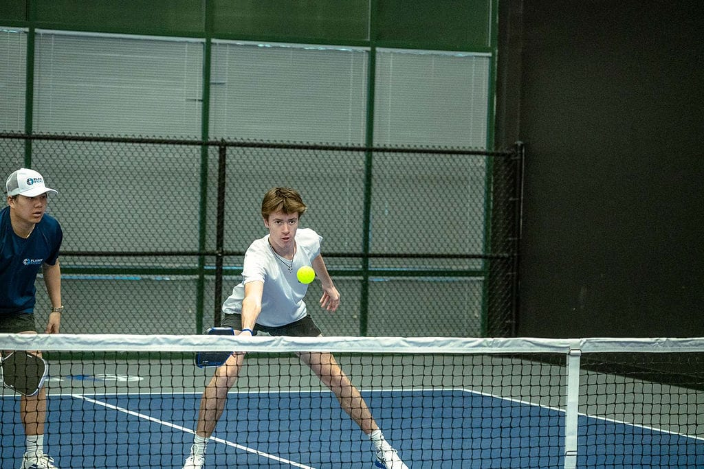 Two people play doubles pickleball indoors; one player prepares to hit the ball with a paddle near the net while the other stands ready behind him.