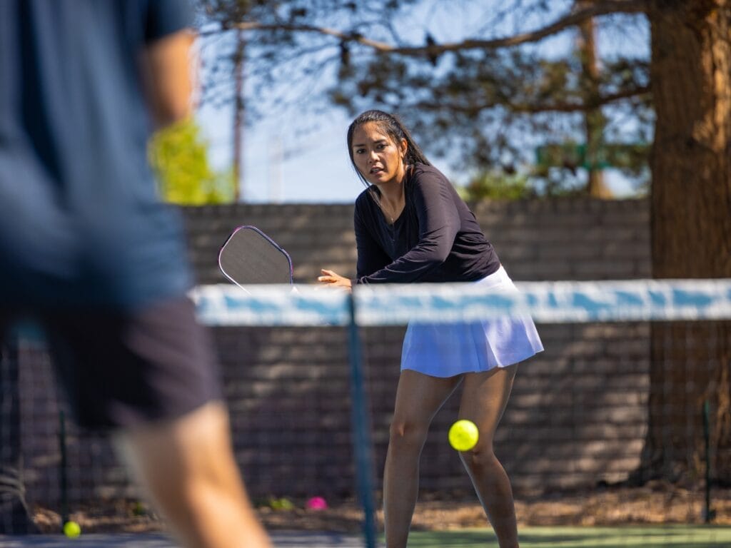 A woman in athletic attire prepares to hit a pickleball during a game on an outdoor court, showcasing her essential pre game checklist skills as a blurred opponent waits in the foreground.