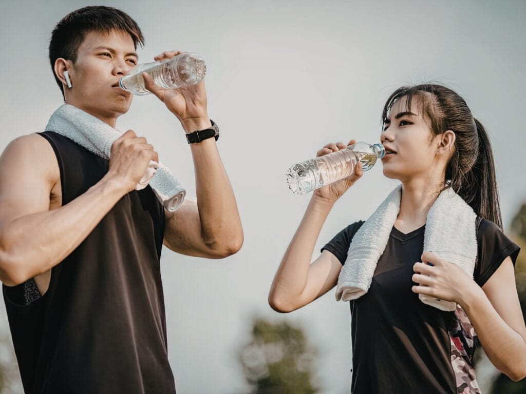 Two people wearing black athletic wear and towels around their necks drink water from bottles outdoors, likely reviewing their pickleball checklist before starting a game.