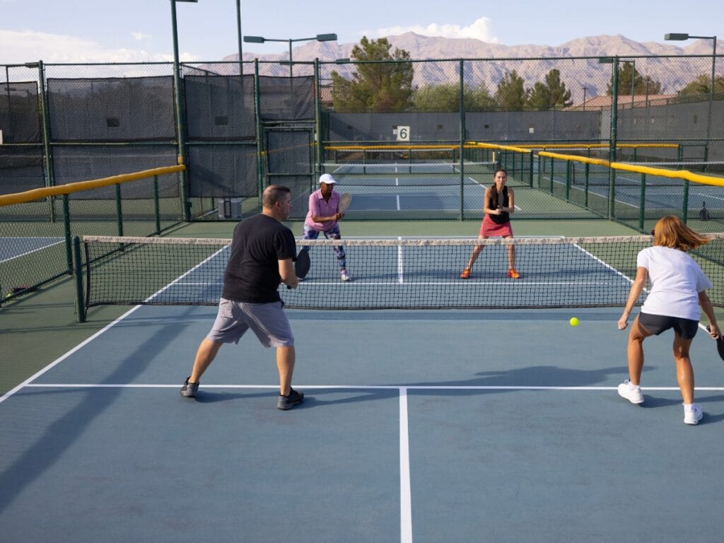 Four people enjoy an essential game of doubles pickleball on an outdoor court, with two players on each side of the net, surrounded by fencing and mountains in the background.