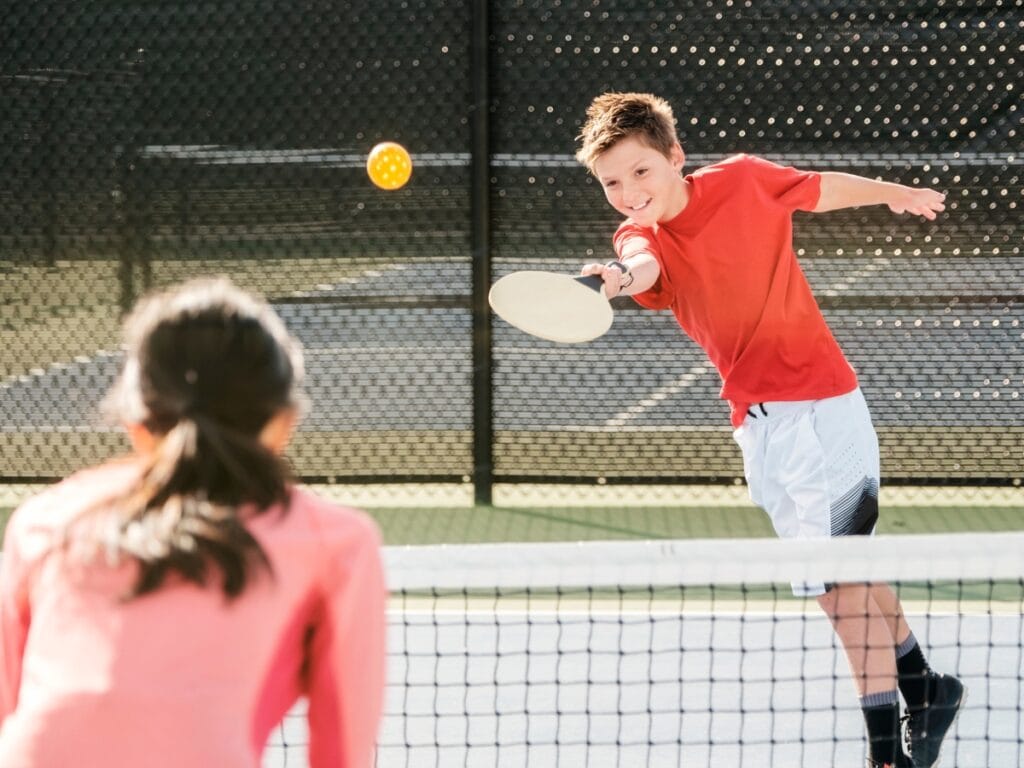 Two children play essential Pickleball on an outdoor court, with one hitting the ball over the net toward the other.