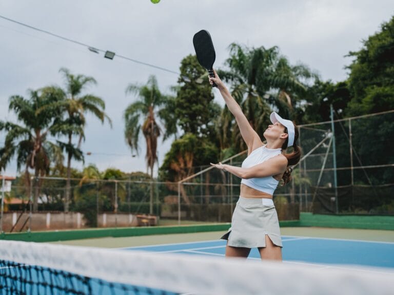 A woman in athletic attire completes her essential pre game checklist as she prepares to hit a pickleball with a paddle on an outdoor court surrounded by palm trees and a fence.