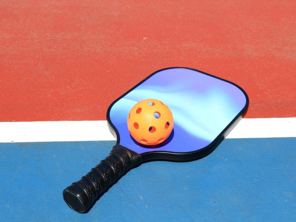 A blue pickleball paddle and an orange perforated ball rest on a court with red, white, and blue surface sections, reminding players that proper paddle care helps keep your equipment game-ready.