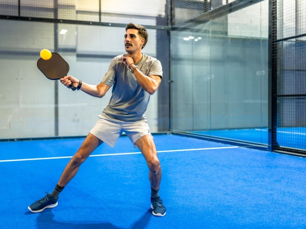 A man in sportswear prepares to hit a ball with a paddle on an indoor blue court enclosed by glass walls, showcasing his skills and dedication to paddle care.