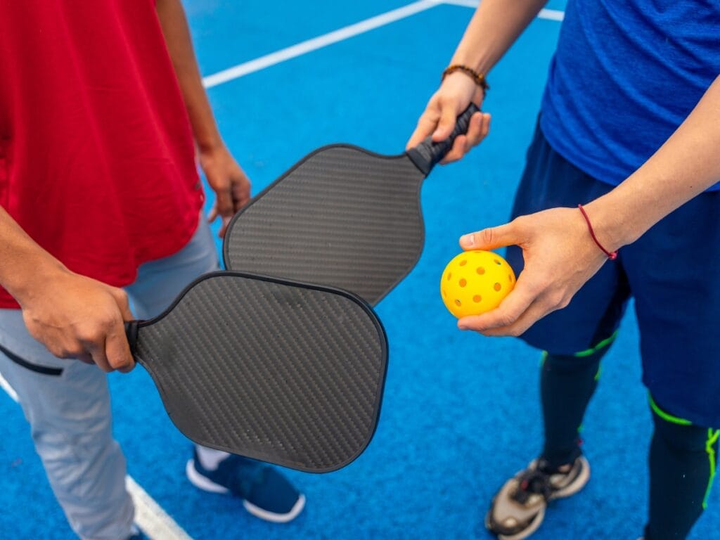 Two people stand on a blue court holding pickleball paddles, with one person holding a yellow perforated ball, ready to share paddle care tips for the perfect game.