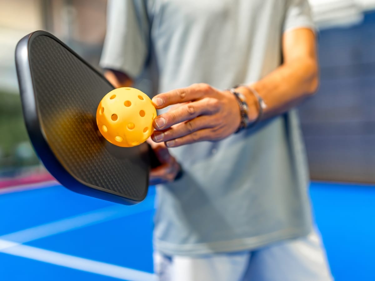 A person holding a pickleball paddle and balancing a yellow perforated pickleball on it, with an indoor court in the background&mdash;demonstrating good paddle care and handling skills.