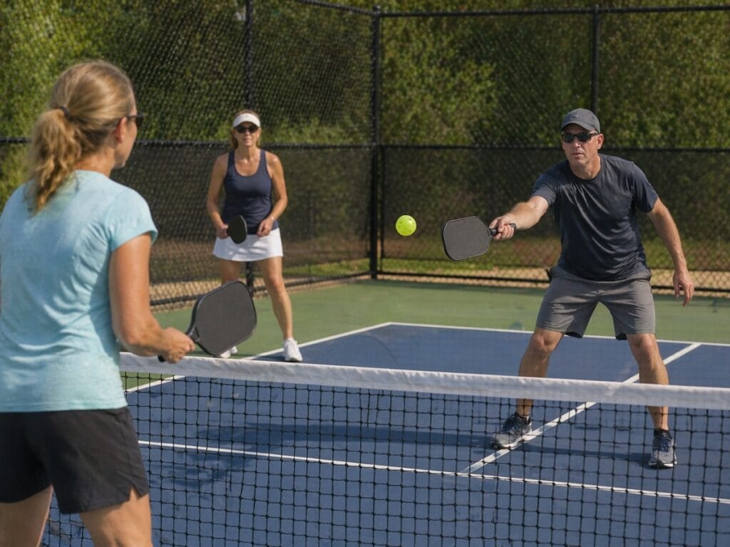 Three adults play pickleball on an outdoor court, with one man hitting the ball over the net toward a woman on the opposite side as they reference a pickleball guide to keep track of scoring.
