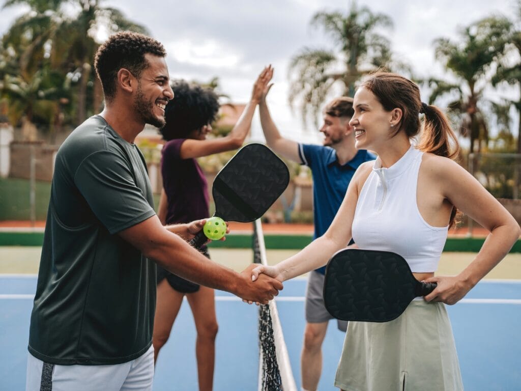 Two pickleball players shake hands at the net while their partners high-five in the background, showing true sportsmanship and teamwork&mdash;essential steps in mastering pickleball on an outdoor court.