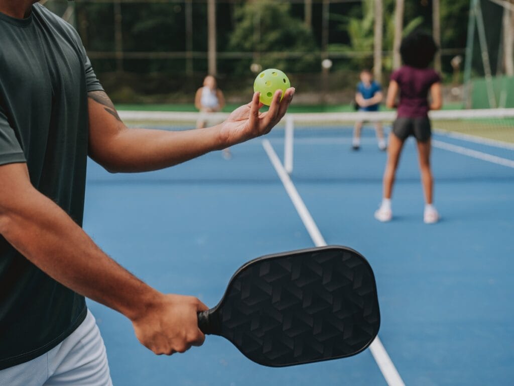 A person prepares to serve with a paddle and yellow plastic ball on a blue outdoor pickleball court, while others stand on the opposite side of the net&mdash;an essential moment in mastering pickleball.