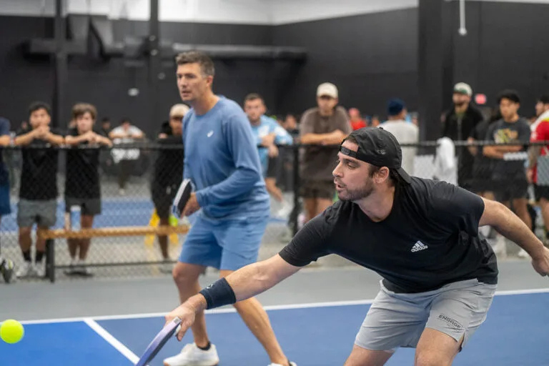 Two men play doubles pickleball indoors while spectators watch from behind a fence; one man is reaching forward to hit the ball with his paddle, showcasing skills from a pickleball guide on mastering pickleball techniques.