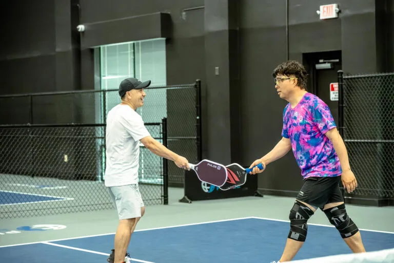 Two men on an indoor pickleball court tap paddles at the net, one in a white outfit and the other in a pink and blue tie-dye shirt with knee braces, exchanging paddle tips for better paddle care.