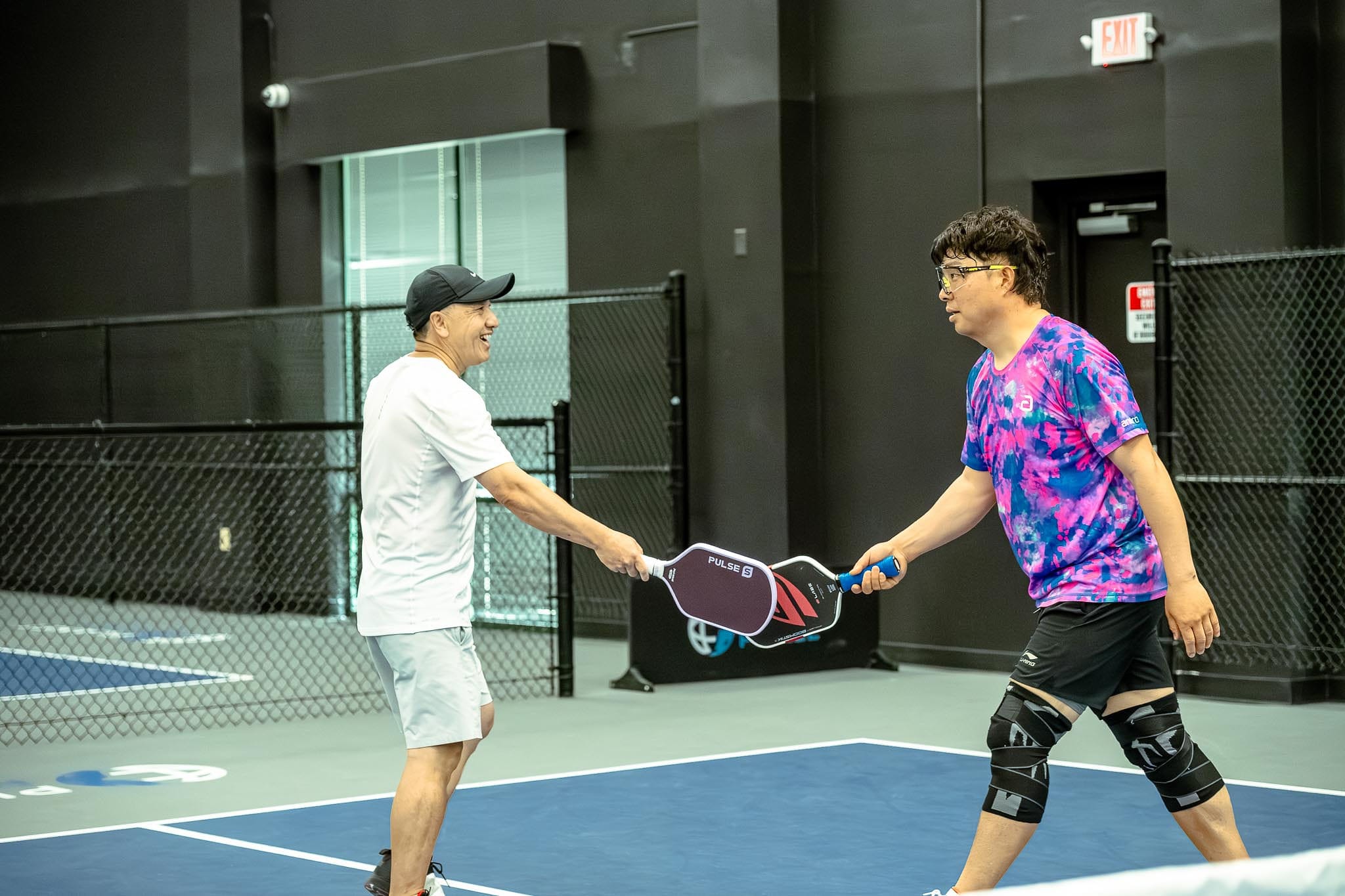 Two men on an indoor pickleball court tap paddles at the net, one in a white outfit and the other in a pink and blue tie-dye shirt with knee braces, exchanging paddle tips for better paddle care.